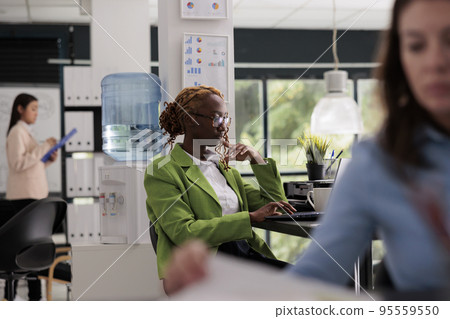 Thoughtful employee working business office open space, manager sitting at workplace desk, colleagues on background. Young woman thinking, typing on computer, side view medium shot 95559550