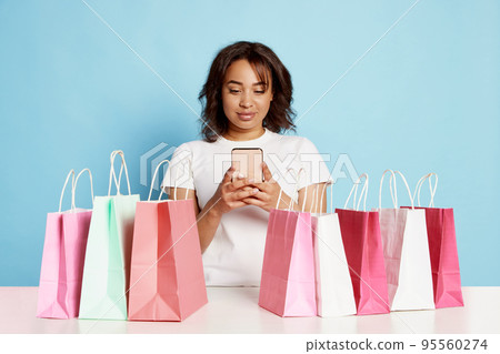 Portrait of beautiful young woman doing online shopping on phone around many shopping bags over blue background 95560274
