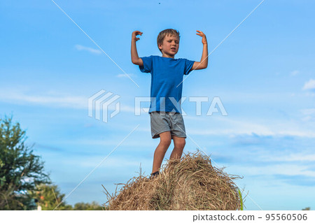 Boy smile play dance grimace show off blue t-shirt stand on haystack bales of dry grass, clear sky sunny day. Balance training. Concept happy childhood, children outdoors, clean air close to nature 95560506