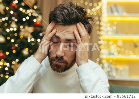 A man has a headache for Christmas, a close-up photo on New Year's Eve near the tree, a man holding his head with his hands, sick, sitting on the sofa at home alone. 95561712