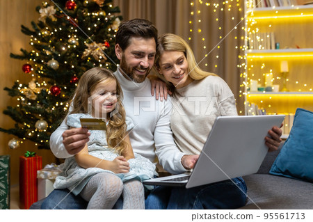 Christmas family, husband wife and little daughter for Christmas at home make online shopping in online store remotely, smiling and happy, husband holding bank credit card and laptop choosing gifts. 95561713