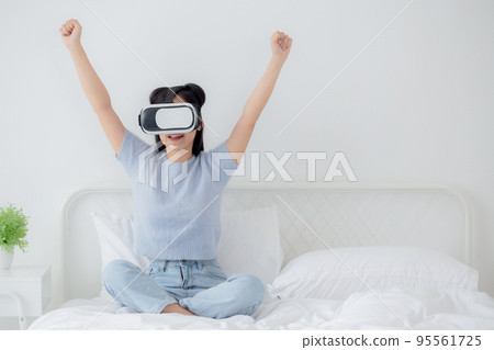 Young asian woman wearing vr headset playing game with excited in the bedroom at home. 95561725