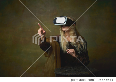 Beautiful young girl with long blonde hair wearing medieval dress and VR headset over dark vintage background. Retro style, art, fashion, comparison of eras concept. 95563654