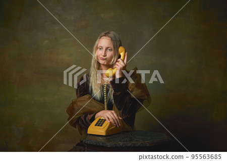 Annoyed young girl with long blonde hair wearing medieval dress like Mona Lisa replica over dark vintage background. Vintage style, art, fashion, comparison of eras concept. Annoyed young girl with long blonde hair wearing medieval dress like Mona Lisa replica over dark vintage background. Vintage style, art, fashion, comparison of eras concept. 95563685