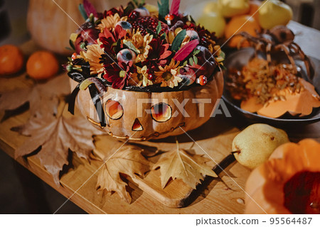 Halloween pumpkin decorated with flowers and gummy eyes on a wooden table. Conceptual still life on the theme of Halloween. 95564487