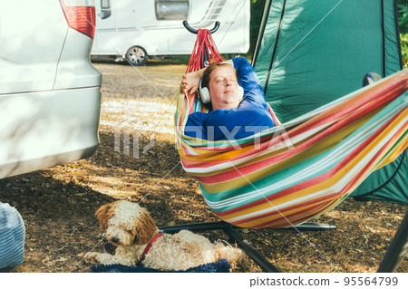 Relaxed woman listening to music with headphones lying in hammock with cockapoo puppy near motorhome on camping trip. Female living on camper car with animal, travel the world. Caravan car Vacation Relaxed woman listening to music with headphones lying in hammock with cockapoo puppy near motorhome on camping trip. Female living on camper car with animal, travel the world. Caravan car Vacation 95564799