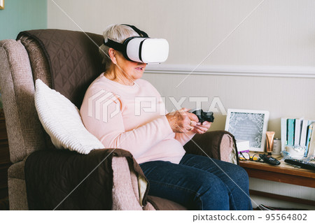 Emotional senior woman wearing virtual reality goggles and using wireless controller, joystick gamepad playing video games at home. Fun on Retirement. Gaming hobby. Technology, entertainment industry. 95564802