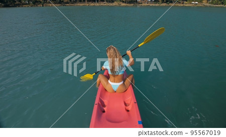 Seductive blonde woman in blue swimsuit sails on pink plastic kayak along azure sea at exotic resort backside upper above view. Seductive blonde woman in blue swimsuit sails on pink plastic kayak along azure sea at exotic resort backside upper above view. 95567019