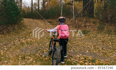 One caucasian children walk with bike in autumn park. Little girl walking black orange cycle in forest. Kid goes do bicycle sports. Biker motion ride with backpack and helmet. Mountain bike hard tail. 95567290