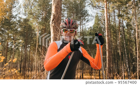 Training an athlete on the roller skaters. Biathlon ride on the roller skis with ski poles, in the helmet. Autumn workout. Roller sport. Athlete is getting ready to start. 95567400