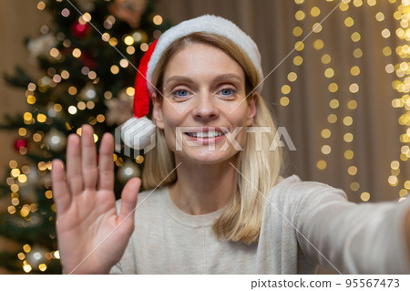 Mature woman at home celebrating New Year and Christmas, blonde talking on video call using app on phone to communicate with friends remotely, wife near Christmas tree in living room at home. 95567473