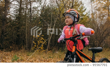 One caucasian children walk with bike in autumn park. Little girl walking black orange cycle in forest. Kid goes do bicycle sports. Biker motion ride with backpack and helmet. Mountain bike hard tail. 95567701
