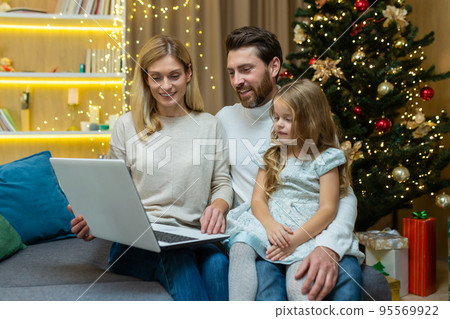 Happy family man wife and daughter celebrating new year and Christmas together, couple sitting on sofa at home near Christmas tree with laptop, watching video online together. Happy family man wife and daughter celebrating new year and Christmas together, couple sitting on sofa at home near Christmas tree with laptop, watching video online together. 95569922