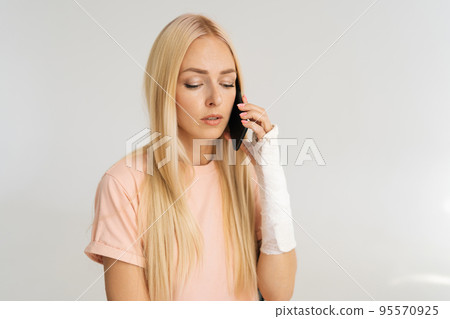 Close-up portrait of worried sad young woman with broken arm wrapped in plaster bandage talking smartphone on white isolated background, looking down. Concept of insurance and healthcare. 95570925