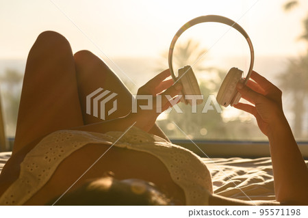Beautiful woman in nightgown with headphones listening music lying on the bed against sea and palm trees at summer sunny day 95571198