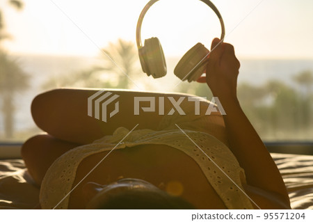Beautiful woman in nightgown with headphones listening music lying on the bed against sea and palm trees at summer sunny day 95571204