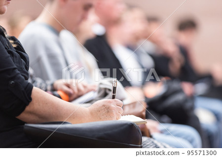 Female hands holding pen and notebook, making notes at conference lecture. Event participants in conference hall. Female hands holding pen and notebook, making notes at conference lecture. Event participants in conference hall. 95571300