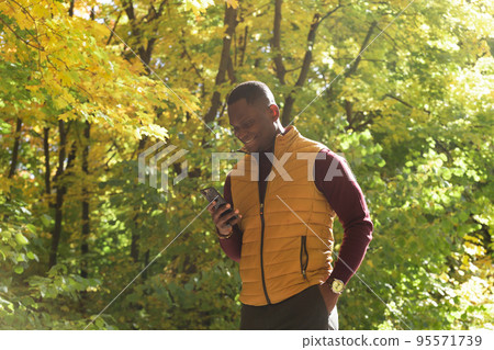 Millennial african american male walking on city street autumn park holding mobile phone for blogging. Smiling man student strolling on urban setting with cellular - social network concept Millennial african american male walking on city street autumn park holding mobile phone for blogging. Smiling man student strolling on urban setting with cellular - social network concept 95571739