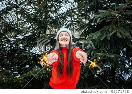 Outdoor portrait of happy cute Latina girl in winter hat and red sweater posing with sparkler against Christmas trees with snow background. Brunette woman having fun in winter holidays. 95571758