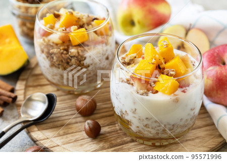Healthy autumn or winter breakfast granola. Homemade dessert with yogurt, granola, caramel apples and pumpkin, cinnamon, nuts on a stone table. 95571996