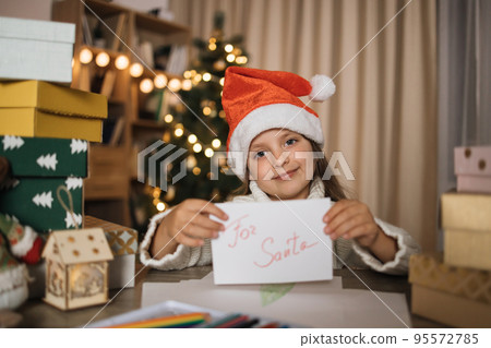 Smiling caucasian girl in red hat holding letter in hands after writing wish list for Santa at home. Smiling caucasian girl in red hat holding letter in hands after writing wish list for Santa at home. 95572785