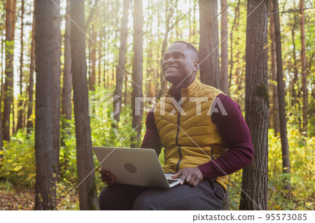African american man sitting in city park on a bench with laptop studying online outdoors. Man freelancer learn working remotely in street e-learning 95573085