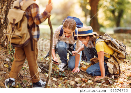 Happy mother and two children in with backpacks examining environment through magnifying glass in forest Happy mother and two children in with backpacks examining environment through magnifying glass in forest 95574753