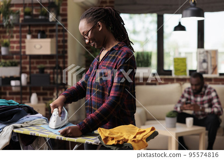 Smiling african american woman ironing clothes at home while distracted husband uses cell phone. Optimistic modern housewife takes care of household chores in urban apartment 95574816