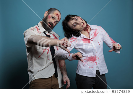 Creepy halloween monsters showing thumbs down on camera, expressing disapproval and dislike in studio. Couple of brain eating zombies with bloody scars and ugly wounds, frightening devils. 95574829