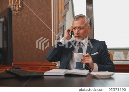 Male hotelier works at a computer and talking phone while sitting in his office in hotel Male hotelier works at a computer and talking phone while sitting in his office in hotel 95575390