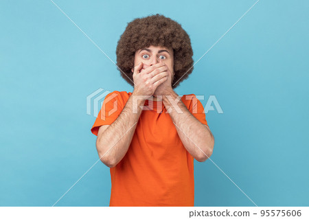 Man with Afro hairstyle in orange T-shirt closing mouth not to scream, feeling frightened and terrified, his eyes and look full of fear and terror. Indoor studio shot isolated on blue background. 95575606