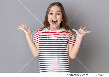 Portrait of excited little girl wearing striped T-shirt standing with raised arms and open mouth, hearing astonishing news, saying wow. Indoor studio shot isolated on gray background. 95575609