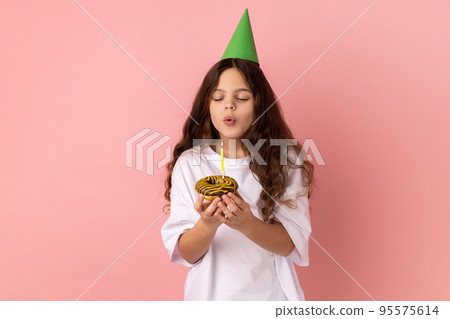 Cute charming little girl in green party cone on head holding cake and blowing candle, making wish, expressing happiness, keeps eyes closed. Indoor studio shot isolated on pink background. Cute charming little girl in green party cone on head holding cake and blowing candle, making wish, expressing happiness, keeps eyes closed. Indoor studio shot isolated on pink background. 95575614