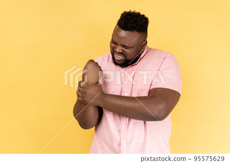 Wrist sprain. Portrait of man wearing pink shirt touching injured hand grimacing in pain, suffering carpal tunnel syndrome, arthritis inflammation. Indoor studio shot isolated on yellow background. Wrist sprain. Portrait of man wearing pink shirt touching injured hand grimacing in pain, suffering carpal tunnel syndrome, arthritis inflammation. Indoor studio shot isolated on yellow background. 95575629