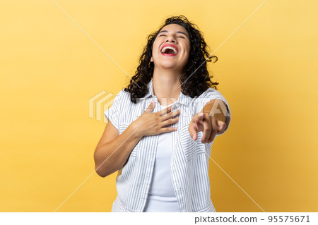 Portrait of happy woman with dark wavy hair laughing out loud and pointing finger to camera, hearing funny joke, holding belly. Indoor studio shot isolated on yellow background. 95575671