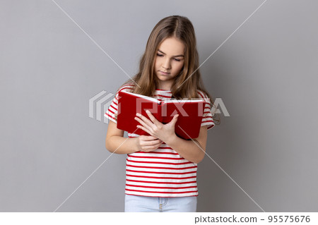 Portrait of concentrated dark haired little girl wearing striped T-shirt holds book, reading, absorbed with interesting plot. Indoor studio shot isolated on gray background. Portrait of concentrated dark haired little girl wearing striped T-shirt holds book, reading, absorbed with interesting plot. Indoor studio shot isolated on gray background. 95575676