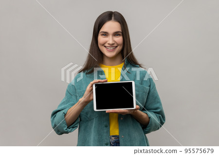 Young smiling woman holding digital tablet in hands, showing blank device screen to camera, expressing happiness, wearing casual style jacket. Indoor studio shot isolated on gray background. Young smiling woman holding digital tablet in hands, showing blank device screen to camera, expressing happiness, wearing casual style jacket. Indoor studio shot isolated on gray background. 95575679