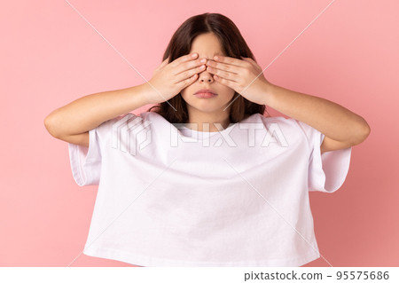Portrait of sad upset little girl wearing white T-shirt standing covering eyes with hand, does not want see at something shameful. Indoor studio shot isolated on pink background. 95575686