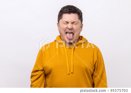 Naughty infantile middle aged man closing eyes and showing tongue, having fun, fooling around, childish manners, wearing urban style hoodie. Indoor studio shot isolated on white background. 95575703