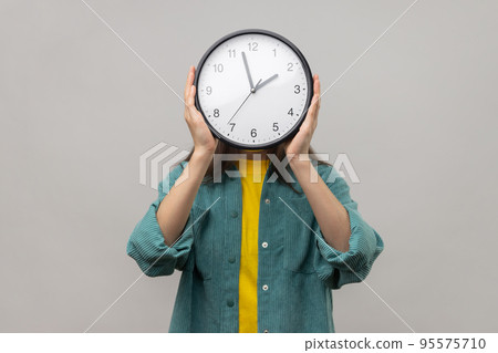 Portrait of unknown woman holding wall clock hiding her face, time management, schedule and meeting appointment, wearing casual style jacket. Indoor studio shot isolated on gray background. 95575710