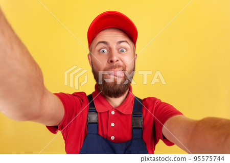 Childish worker man wearing red cap and blue uniform taking selfie, looking at camera and showing tongue out POV, point of view of photo. Indoor studio shot isolated on yellow background. Childish worker man wearing red cap and blue uniform taking selfie, looking at camera and showing tongue out POV, point of view of photo. Indoor studio shot isolated on yellow background. 95575744