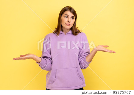 I don't know. Portrait of confused beautiful young woman standing with raised arms and looking at camera, wearing purple hoodie. Indoor studio shot isolated on yellow background. 95575767