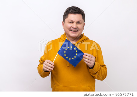 European Union flag. Portrait of happy man with dark hair smiling and holding EU flag, looking at camera with toothy smile, wearing urban style hoodie. Indoor studio shot isolated on white background. 95575771