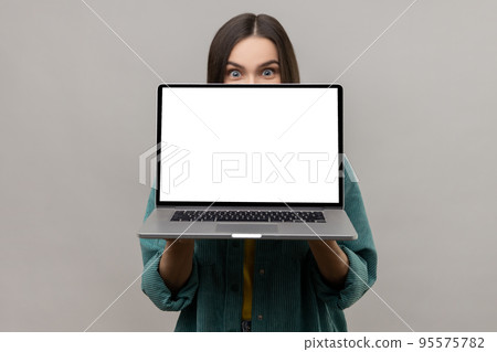 Positive unknown woman hiding half of face behind laptop with white empty display, looking at camera with big surprised eyes, wearing casual jacket. Indoor studio shot isolated on gray background. 95575782