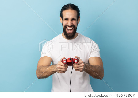 Portrait of man with beard wearing white T-shirt holding in hands red gamepad joystick, looking at camera with optimistic expression, enjoying game. Indoor studio shot isolated on blue background. Portrait of man with beard wearing white T-shirt holding in hands red gamepad joystick, looking at camera with optimistic expression, enjoying game. Indoor studio shot isolated on blue background. 95575807