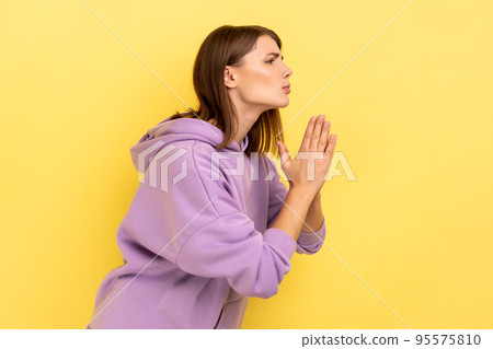 Side view of pleading woman keeping arms in prayer gesture and asking forgiveness, feeling sorry for mistake, wearing purple hoodie. Indoor studio shot isolated on yellow background. Side view of pleading woman keeping arms in prayer gesture and asking forgiveness, feeling sorry for mistake, wearing purple hoodie. Indoor studio shot isolated on yellow background. 95575810