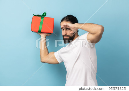 Portrait of man with beard wearing white T-shirt holding gift box, looks far away with hand over eyes, waits future holidays, birthday celebration. Indoor studio shot isolated on blue background. Portrait of man with beard wearing white T-shirt holding gift box, looks far away with hand over eyes, waits future holidays, birthday celebration. Indoor studio shot isolated on blue background. 95575825