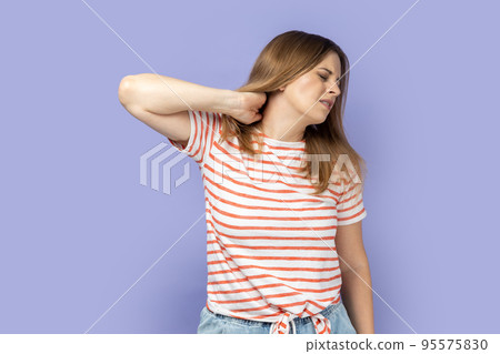 Portrait of attractive tired upset unhealthy blond woman wearing striped T-shirt standing massaging neck to relieve pain, muscle strain in back. Indoor studio shot isolated on purple background. 95575830