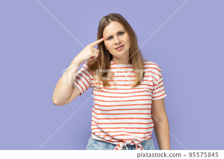 Unhappy frustrated blond woman wearing striped T-shirt showing stupid gesture, does not believe in his strengths, capabilities, disappointed. Indoor studio shot isolated on purple background. Unhappy frustrated blond woman wearing striped T-shirt showing stupid gesture, does not believe in his strengths, capabilities, disappointed. Indoor studio shot isolated on purple background. 95575845