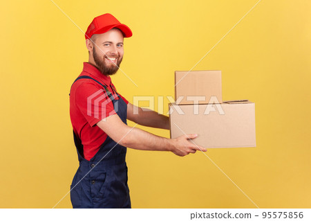 Side view of happy deliveryman holding two cardboard boxes, delivering order door-to-door, shipment and cargo transportation service. Indoor studio shot isolated on yellow background. Side view of happy deliveryman holding two cardboard boxes, delivering order door-to-door, shipment and cargo transportation service. Indoor studio shot isolated on yellow background. 95575856